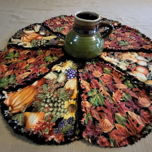 Könnte beinhalten: Ein handgefertigter, kreisförmiger Tischset mit schwarzem Rand und einem herbstlichen Stoff mit Blättern, Kürbissen, Trauben und Sonnenblumen. Eine grüne Keramiktasse mit braunem Rand und Henkel steht in der Mitte des Sets.