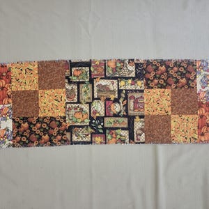 May include: A rectangular fabric table runner featuring a patchwork design of various autumn-themed patterns. The print includes illustrations of orange pumpkins, sunflowers, red barns, and corn stalks in shades of orange, brown, yellow, and black.