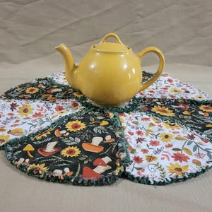 May include: A round, quilted fabric mat featuring alternating panels of black fabric with a mushroom and sunflower print and white fabric with a floral and leaf pattern, topped with a solid yellow ceramic teapot with a curved handle and spout.