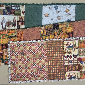 May include: A rectangular rag quilt table runner featuring a patchwork design with autumn-themed fabrics, including pumpkins, sunflowers, birds, and figures with the text Give Thanks. The edges are finished with a frayed, white and green fringe.