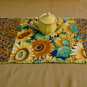 May include: A yellow and blue table runner with a sunflower pattern. A yellow ceramic sugar bowl sits in the center of the runner.
