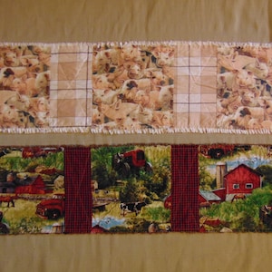 May include: Two quilted table runners, one with a brown and white checked pattern and a piglet print, and the other with a red and black checked pattern and a farm scene with a red barn.