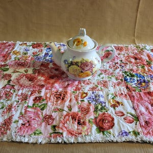 May include: A white teapot with a floral design sits on a white tablecloth with a patchwork of pink and white floral patterns.
