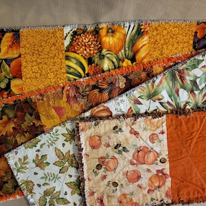 May include: A patchwork table runner with autumnal patterns. The runner displays fabrics with pumpkins, falling leaves, and floral designs in orange, yellow, and green hues. The edges have a fringed trim, giving it a rustic appearance. The runner is approximately 150 cm long.