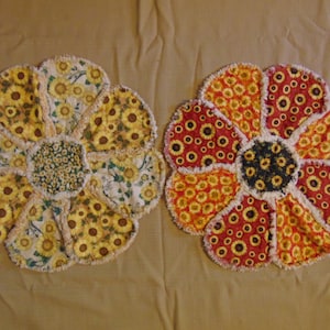 Op de afbeelding: Twee handgemaakte bloemvormige onderzetters van stof. De ene onderzetter is geel en wit met een zonnebloemmotief. De andere onderzetter is rood en oranje met een zonnebloemmotief.