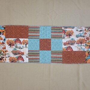 May include: A rectangular fabric table runner featuring a patchwork design of various autumn-themed patterns. The print includes orange pumpkins, red barns, vintage trucks, brown floral motifs, teal swirls, and multi-coloured horizontal stripes.