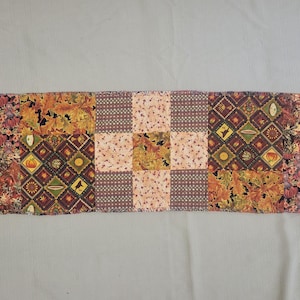 May include: A rectangular patchwork table runner featuring a variety of autumn-themed fabric squares. The design includes patterns of pumpkins, fall leaves, and geometric shapes in shades of orange, brown, black, and cream, arranged in a grid.