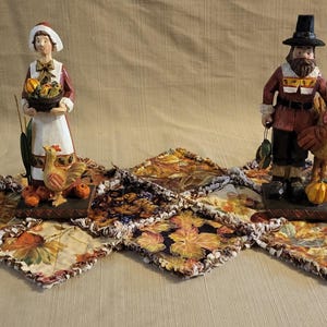 May include: Thanksgiving-themed figurines of a woman and a man, each holding items associated with the holiday. The woman holds a basket of produce, and the man holds a turkey. They stand on a decorative leaf-patterned runner.