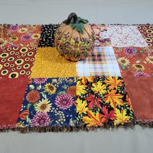Peut inclure: Un chemin de table patchwork rectangulaire composé de carrés de tissu aux motifs automnaux de tournesols, de feuilles d'érable et de fleurs dans des tons orange, jaune et marron, surmonté d'une citrouille décorative en céramique.