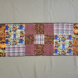 May include: A rectangular rag quilt table runner featuring a patchwork design of autumn-themed fabrics. The pattern includes squares with scarecrows, pumpkins, sunflowers, plaid, and solid maroon textures, all finished with frayed fabric edges.