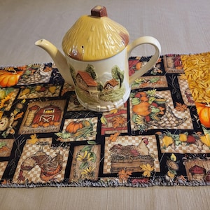 May include: A cream-coloured ceramic teapot with a yellow thatched roof lid rests on a patchwork table runner. The runner displays autumn-themed squares with pumpkins, sunflowers, and farm scenes. The teapot has a rural landscape design.