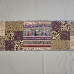 May include: A rectangular fabric table runner featuring a patchwork design with various earth-toned floral and leaf patterns. The centre panel displays a horizontal row of stylised figures holding hands, interspersed with blue and red decorative stripes.