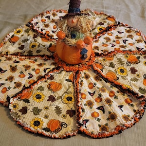 Könnte beinhalten: Herbstliche Tischdecke in Kürbisform. Der cremefarbene Hintergrund ist mit orangefarbenen Kürbissen, Sonnenblumen und Herbstblättern bedruckt. Eine Vogelscheuchenfigur sitzt auf dem Kürbis und verleiht der Herbstdekoration eine festliche Note.
