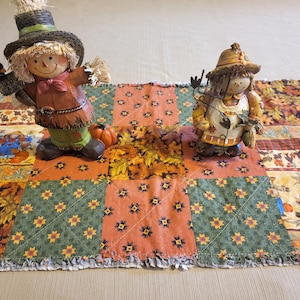 May include: Two decorative scarecrow figurines on a patchwork table runner. The scarecrows wear hats and autumn-themed clothing. The table runner features orange, green, and brown squares with leaf and floral patterns. A small pumpkin sits near one scarecrow.