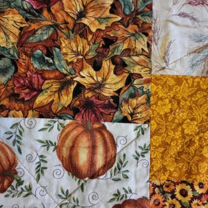May include: A patchwork quilt with autumnal patterns. The quilt displays panels with colourful autumn leaves, pumpkins, floral designs, and sunflowers. The colours include orange, yellow, brown, and green, creating a cosy, seasonal atmosphere.