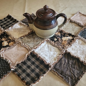 May include: A brown and cream-colored ceramic teapot with a lid sits on a patchwork quilted table runner. The runner features a variety of patterned fabrics in neutral tones, including floral, plaid, and solid squares, with a ruffled edge.