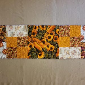 May include: A rectangular patchwork table runner with autumn patterns. The design includes sunflowers, corn, pumpkins, and fall leaves in yellow, orange, and brown. The edges have a frayed, unfinished appearance.