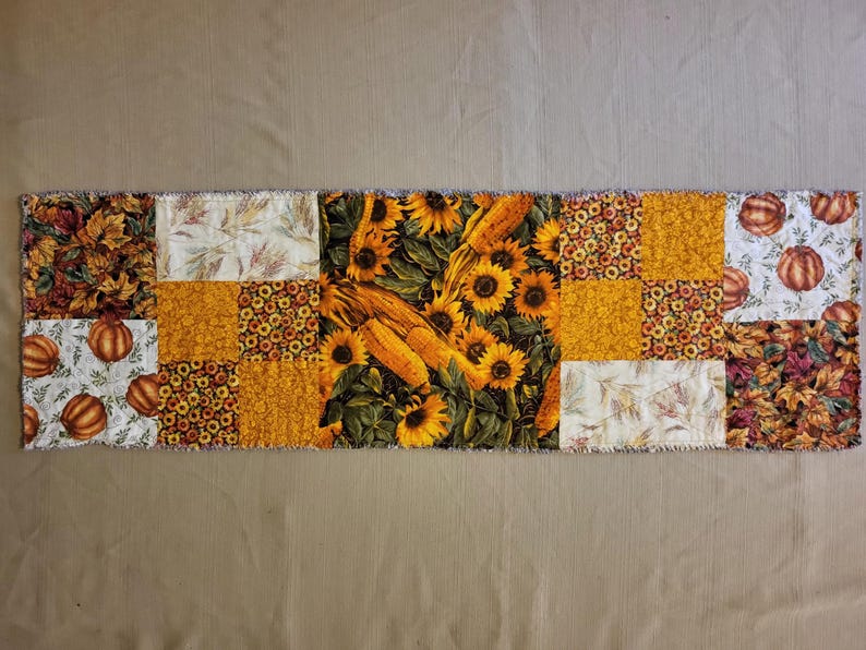 May include: A rectangular patchwork table runner with autumn patterns. The design includes sunflowers, corn, pumpkins, and fall leaves in yellow, orange, and brown. The edges have a frayed, unfinished appearance.