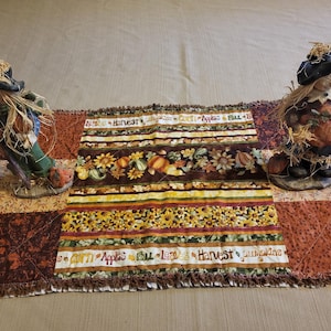 May include: Two scarecrow figurines either side of a fall-themed table runner. The runner has stripes of sunflowers, pumpkins, and the words "Harvest", "Apples", and "Fall". The scarecrows wear hats and hold pumpkins, with straw hair.