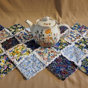 May include: A white porcelain teapot with floral designs sits on a colorful patchwork quilt. The quilt features a variety of floral patterns in shades of blue, yellow, and pink. The teapot has a gold trim and a matching lid.