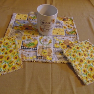 May include: A white mug with the text "HOME is where our story begins" sits on a placemat and two matching coasters. The placemat and coasters feature a patchwork design with sunflowers, honeycombs, and bees.