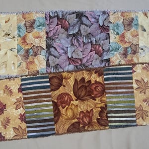 May include: Two rectangular fabric table runners featuring patchwork designs. The top runner displays yellow, orange, and purple leaf patterns. The bottom runner features brown floral motifs, horizontal stripes, and yellow leaf prints with frayed edges.