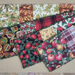 Puede incluir: Un camino de mesa tipo rag quilt rectangular que presenta un mosaico de cuadrados de tela con estampados de temática otoñal, incluyendo manzanas rojas, hojas verdes y marrones, girasoles y patrones de cuadros rojos y blancos, unidos con costuras verdes deshilachadas.