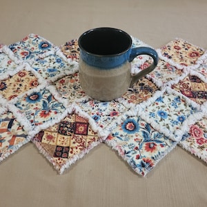 May include: A blue and tan ceramic mug sits on a floral patchwork table runner. The runner features a repeating pattern of colorful floral squares with a white fringe edge. The mug has a blue rim and handle.