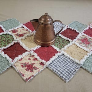 May include: A copper tea kettle rests on a quilted table runner. The runner has a patchwork design with red, green, cream, and patterned fabric squares. The edges of the squares are frayed.