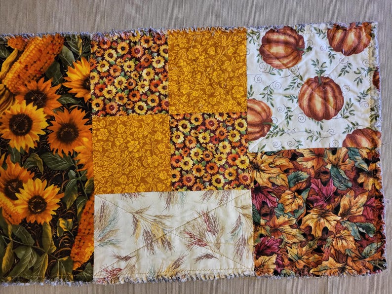 May include: A patchwork quilt with autumn patterns. The quilt displays squares with sunflowers, pumpkins, fall leaves, and wheat stalks in orange, yellow, and brown. The edges have a frayed, textured finish.