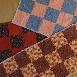 May include: Three patchwork quilt table runners in different patterns and colors. The top runner is pink and blue, the middle runner is red and black, and the bottom runner is brown and beige.