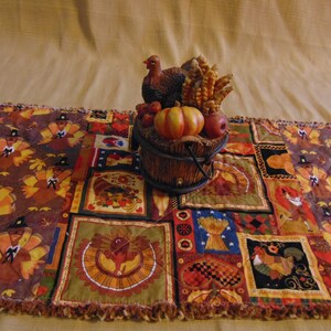 May include: A Thanksgiving-themed table runner with a patchwork design featuring turkeys, pumpkins, and other fall harvest elements. A decorative basket holds a turkey figurine, pumpkins, apples, and wheat stalks. The runner is brown, orange, and yellow.