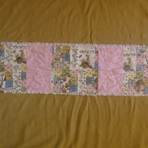 May include: A rectangular table runner with a patchwork design featuring bunnies, carrots, and floral patterns in pastel colours. The centre sections are solid pink, and the edges have a frayed, unfinished look.