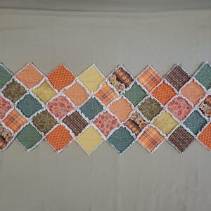 May include: A rectangular table runner composed of diamond-shaped fabric patches in shades of orange, yellow, green, and brown. The patches feature various patterns including floral, plaid, and geometric prints, joined with exposed white frayed seams.