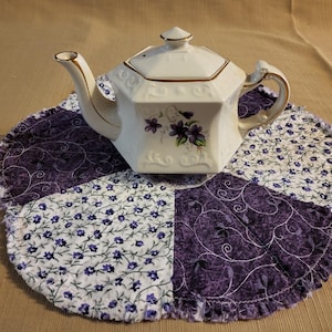 May include: A white teapot with a gold trim and purple flower design sits on a purple and white floral fabric tea cozy.