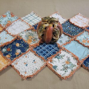 May include: A decorative pumpkin-shaped ceramic candle holder sits on a quilted table runner. The runner features a patchwork design with various floral and patterned fabrics in shades of blue, orange, and yellow. The edges are trimmed with a ruffled border.