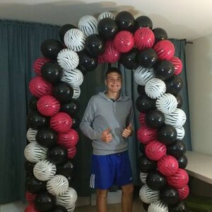 May include: A black, white, and pink balloon archway with a person standing in front of it. The balloons are arranged in a spiral pattern and are tied together with string.