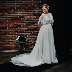 May include: A woman wearing a white lace wedding dress with a tulle skirt. The dress has a lace bodice with a V-neckline and a sash at the waist. The woman is standing in front of a brick wall and a black backdrop.