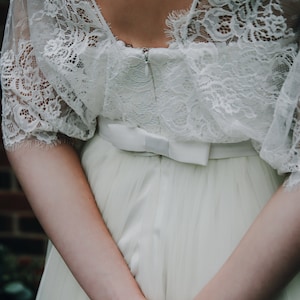 May include: A white lace wedding dress with a satin bow at the waist. The dress has a deep v-neckline and a flowing skirt.