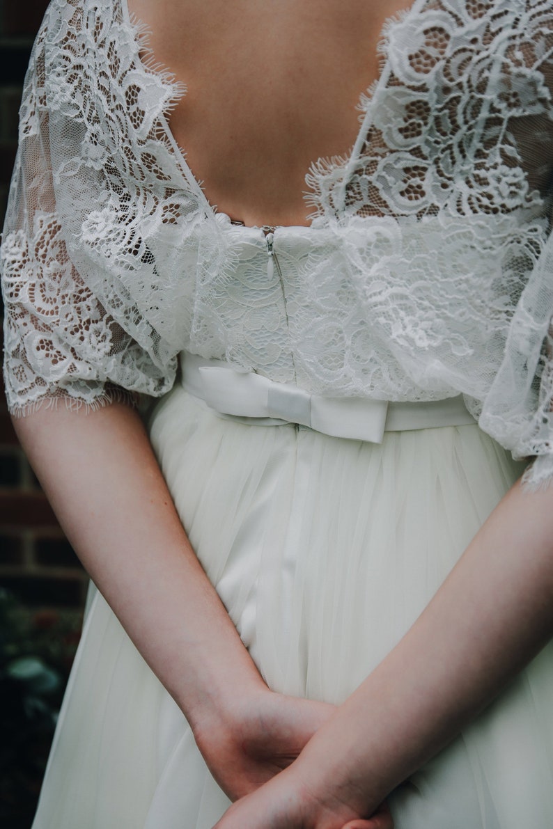 May include: A white lace wedding dress with a satin bow at the waist. The dress has a deep v-neckline and a flowing skirt.