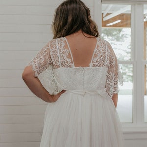 May include: A white lace wedding dress with a tulle skirt and a low back. The dress has a fitted bodice and a flowing skirt. The dress is shown from the back.