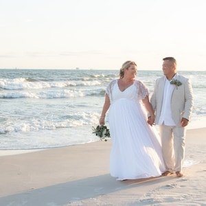 May include: A couple in wedding attire walking on a sandy beach. The bride is wearing a white dress and the groom is wearing a light beige suit. The ocean is in the background with waves crashing on the shore.