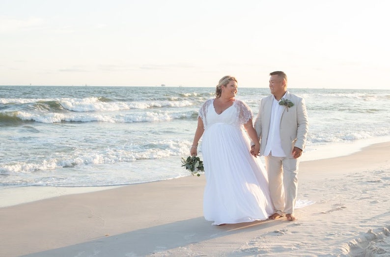 May include: A couple in wedding attire walking on a sandy beach. The bride is wearing a white dress and the groom is wearing a light beige suit. The ocean is in the background with waves crashing on the shore.