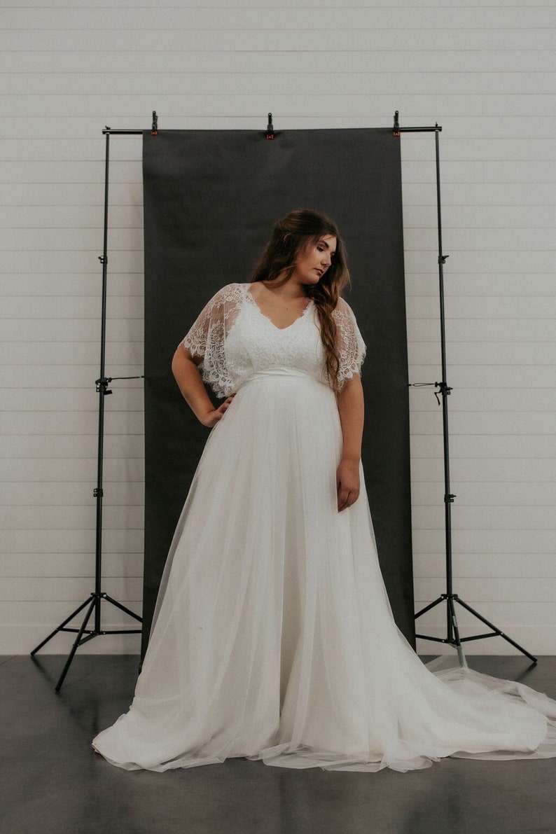 May include: A white lace wedding dress with a tulle skirt and a lace overlay. The dress has a v-neckline and a fitted bodice. The dress is being worn by a model in a studio setting.