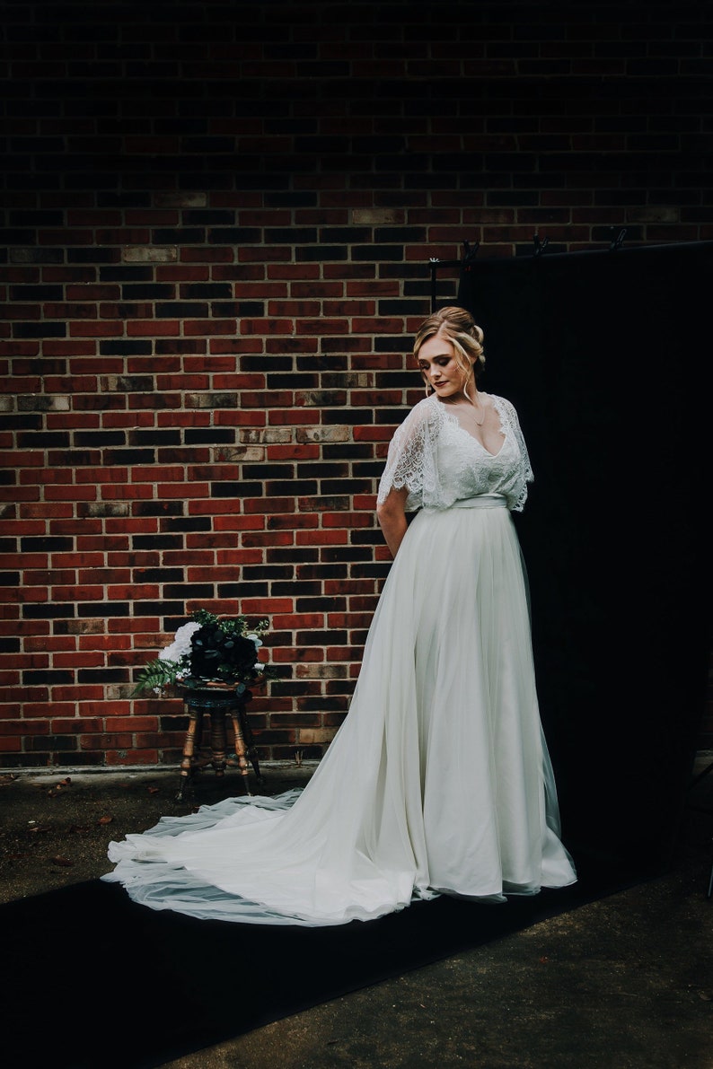May include: A woman wearing a white lace wedding dress with a tulle skirt. The dress has a lace bodice with a V-neckline and a sash at the waist. The woman is standing in front of a brick wall and a black backdrop.