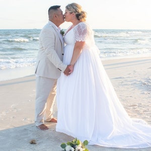 May include: A couple in wedding attire kissing on a beach. The bride is wearing a white lace dress with a long train. The groom is wearing a light beige suit. The couple is standing on a sandy beach with the ocean in the background. A bouquet of white flowers is on the sand in front of the couple.