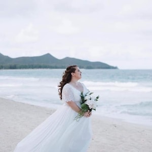 May include: A bride in a white wedding dress with lace sleeves stands on a sandy beach with a bouquet of white flowers in her hands. The bride is looking to the right and smiling. The ocean is in the background.