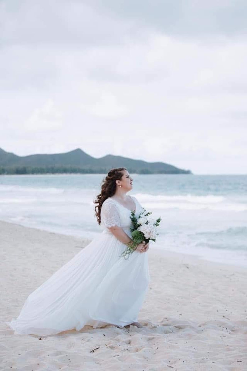 May include: A bride in a white wedding dress with lace sleeves stands on a sandy beach with a bouquet of white flowers in her hands. The bride is looking to the right and smiling. The ocean is in the background.
