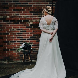 May include: A white lace wedding dress with a tulle skirt. The dress has a low back and a fitted bodice. The dress is displayed on a model against a brick wall and black backdrop.