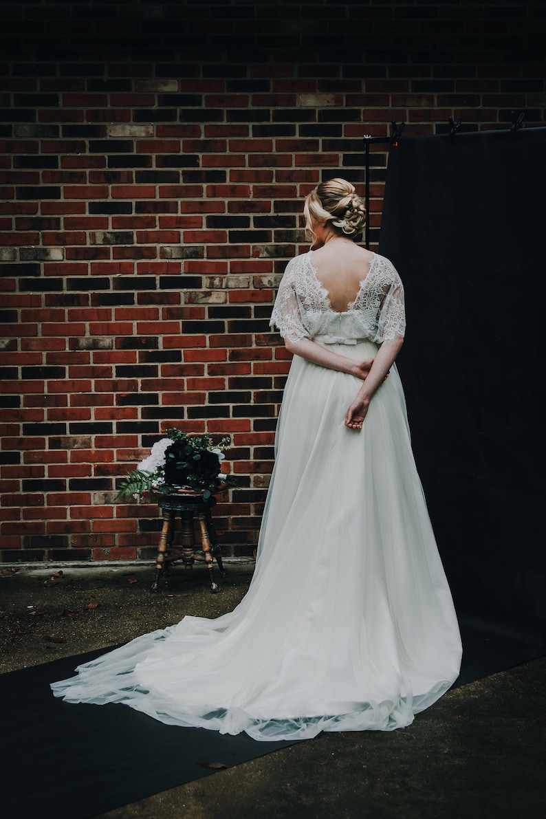 May include: A white lace wedding dress with a tulle skirt. The dress has a low back and a fitted bodice. The dress is displayed on a model against a brick wall and black backdrop.